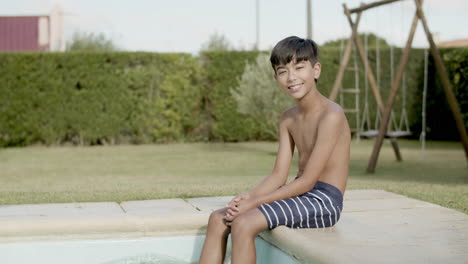 asian boy sitting on pool edge smiling, dangling feet in water.