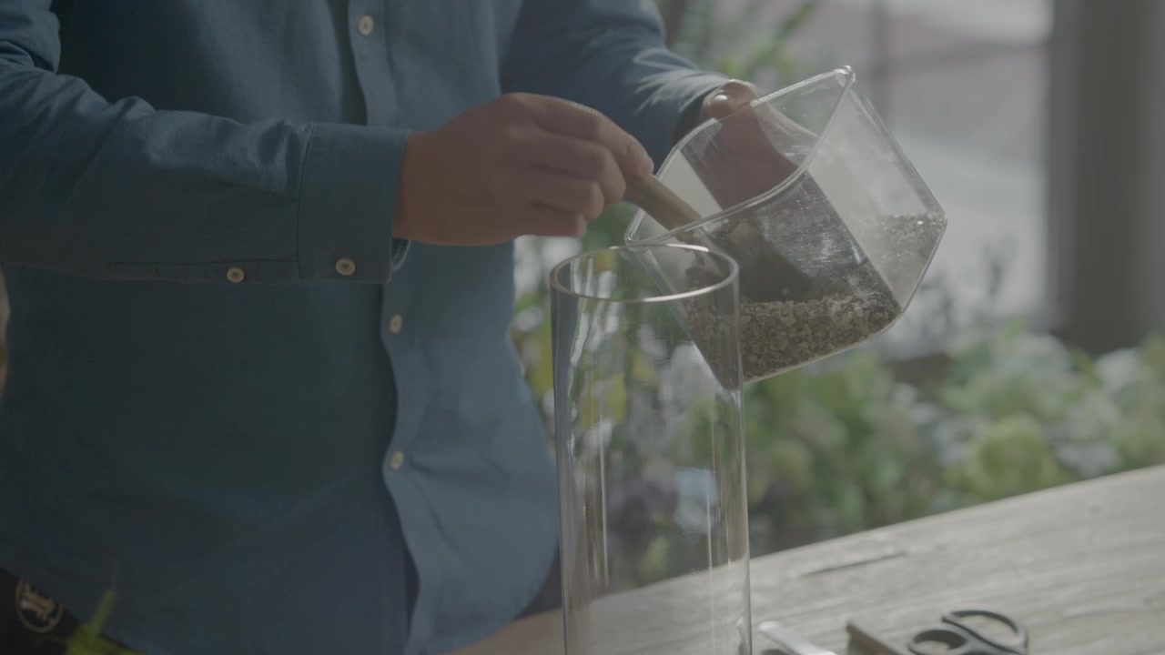 A person carefully pours small pebbles from a clear container into a tall glass vase on a wooden table, surrounded by crafting tools and a natural background.