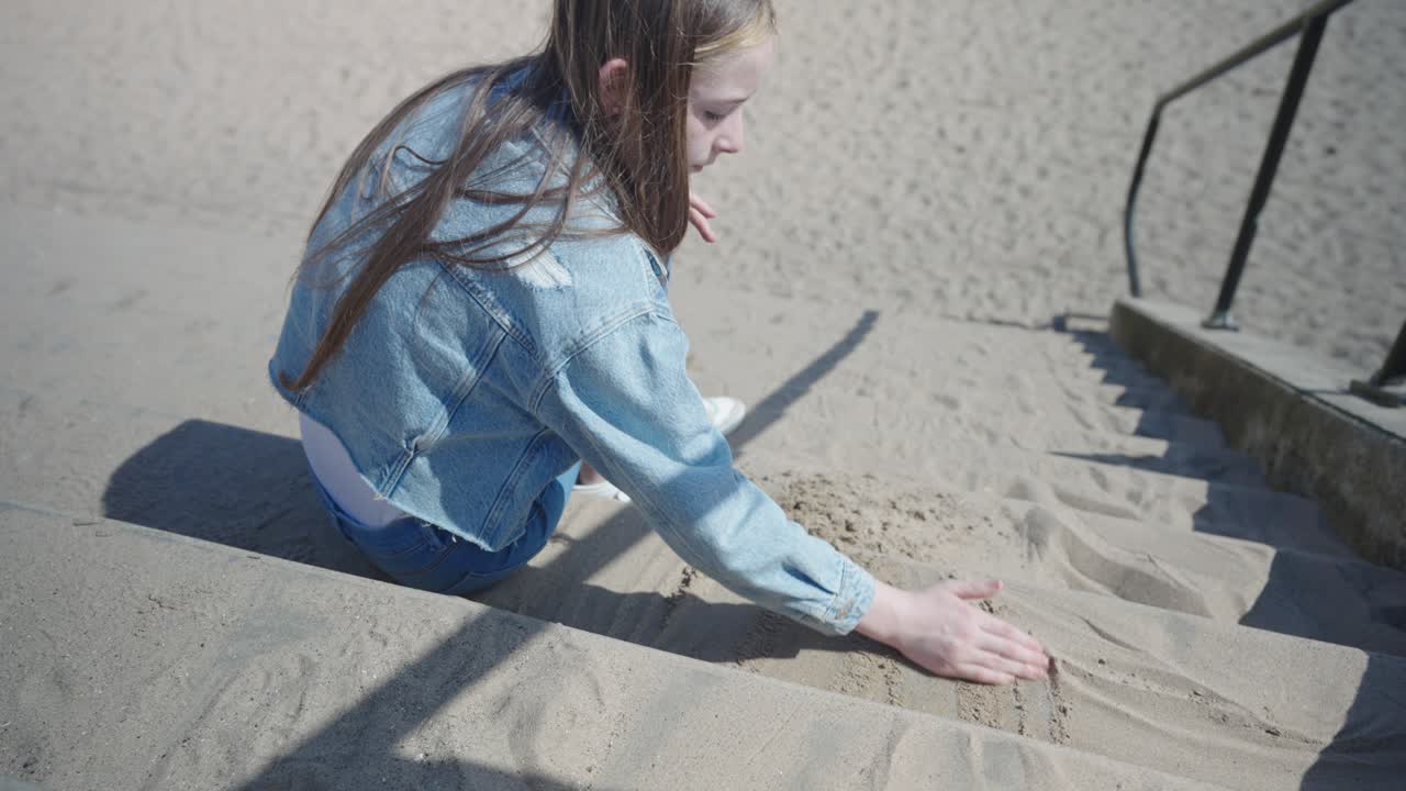 Teenage girl wearing denim playing with sand on steps near beach, medium shot