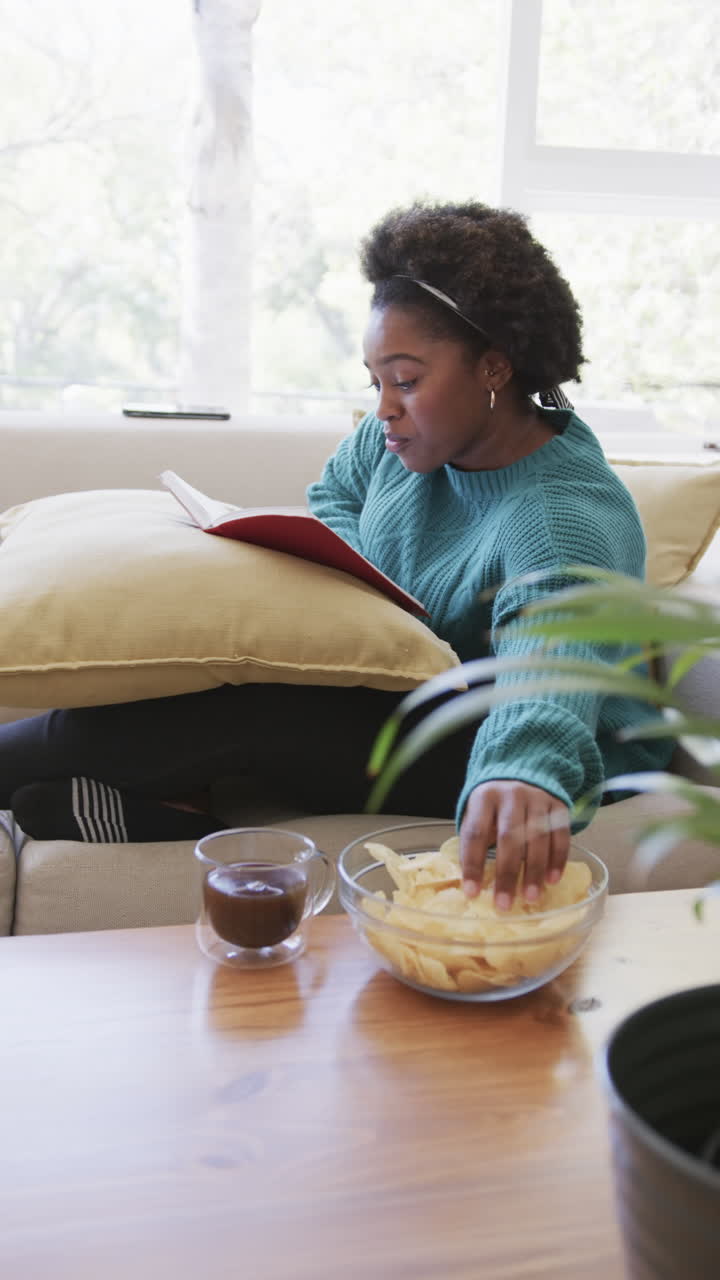 Vertical video of african american woman relaxing and reading book at home
