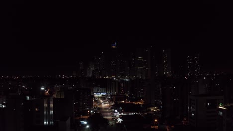 Rising-aerial-drone-night-shot-of-the-beautiful-tropical-beach-capital-city-of-Joao-Pessoa,-Paraiba,-Brazil-from-the-Tambaú-neighborhood-with-skyscrapers,-apartments,-and-a-small-street-with-traffic