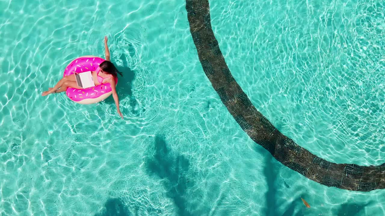 Aerial view of a young brunette woman swimming on an inflatable big donut with a laptop in a transparent turquoise pool