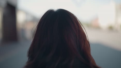 back view of woman with brown hair standing outside in an urban environment, looking away with a soft focus on her hair and surroundings in the background