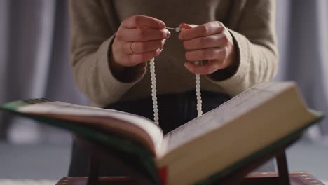 close up of muslim woman praying with prayer beads over open copy of the quran at home