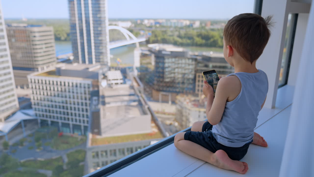 Rear view of a baby boy sitting in front of the panoramic window. Kid takes a picture of the city on the phone camera