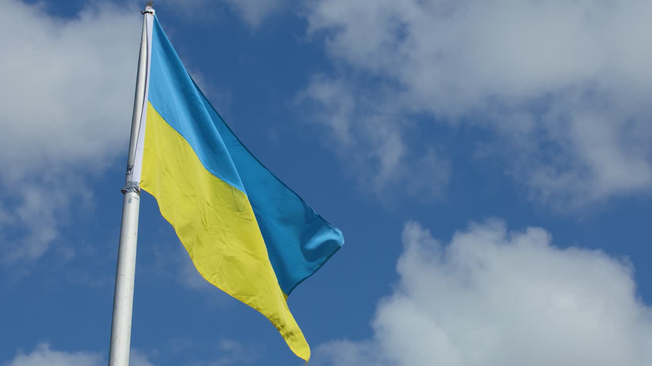Ukrainian flag waves on flagpole, bright daylight, blue sky, scattered clouds, gentle breeze