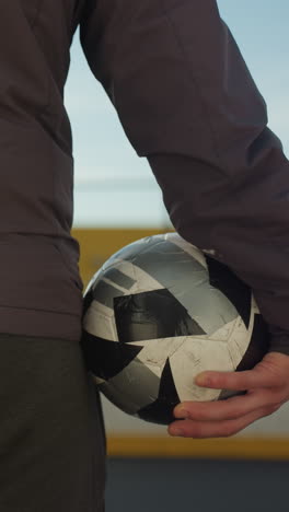 vista trasera de primer plano de un individuo sosteniendo una pelota de fútbol, listo para entrar en la arena deportiva, mostrando ropa atlética y telón de fondo urbano iluminado por la suave luz del sol