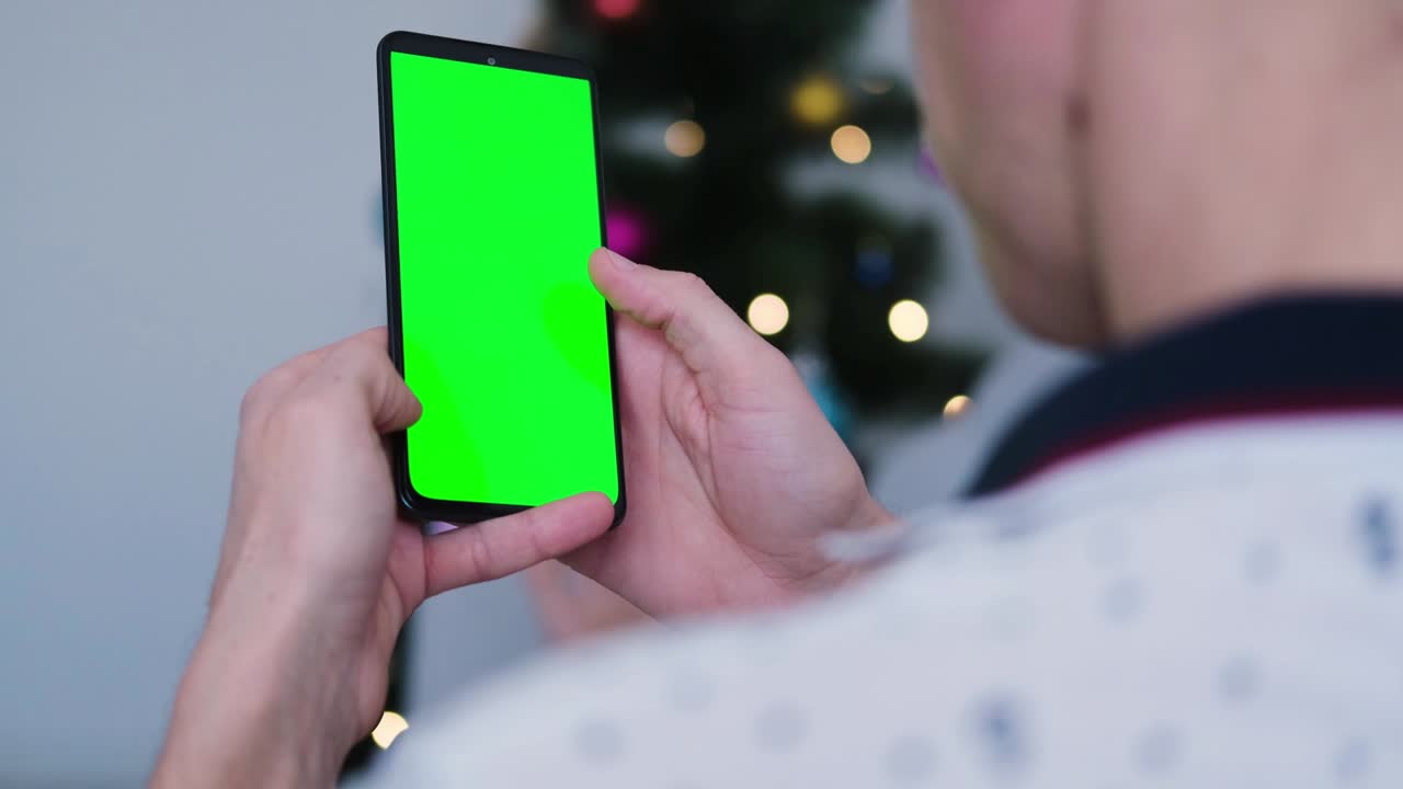 크리스마스 트리를 배경으로 녹색 화면으로 휴대 전화를 사용하는 사람의 손의 클로즈업  - 손가락이 휴대 전화의 녹색 스크린에서 위아래로 스와이프합니다.