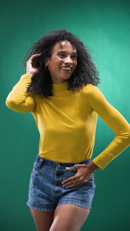 vertical video: confident woman in yellow top adjusting curly hair against green backdrop