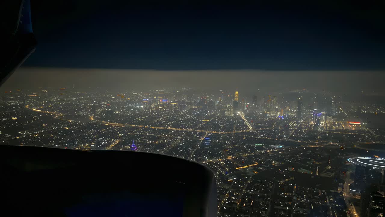 夜、明かりのついた都市のグリッド上を降下する飛行機を録画し、翼と街の明かりを捉えています。