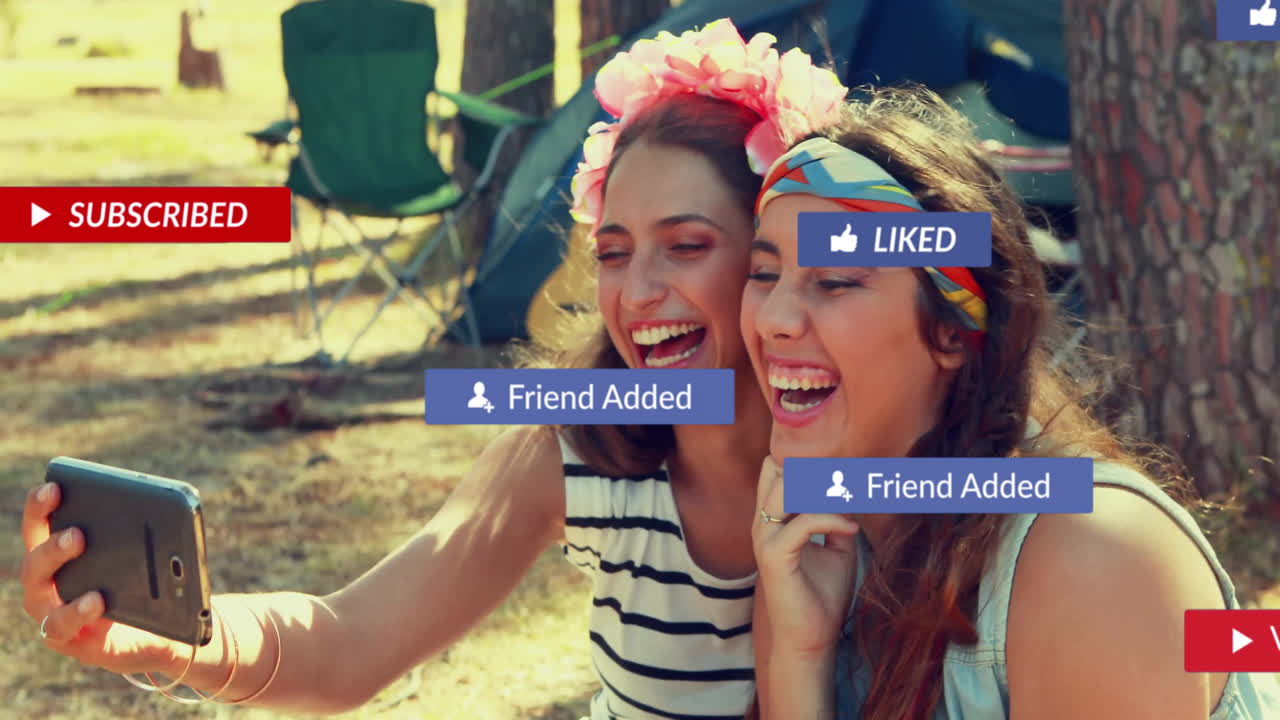 Two friends taking selfie at campsite with tent, using travel app overlay with floating phone icons