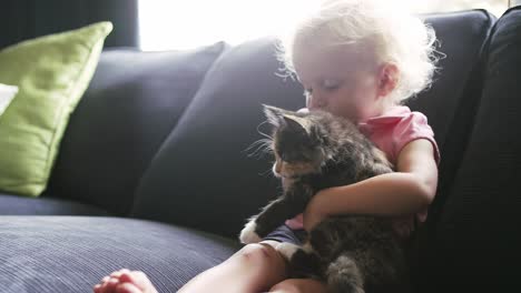 a little girl sitting on the couch holding a kitten and giving it a hug