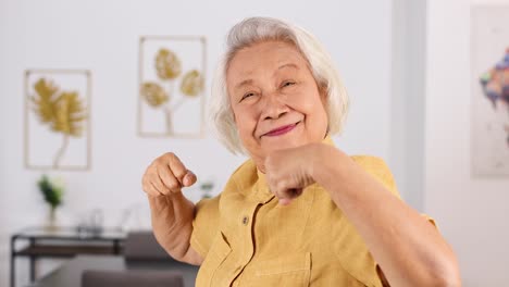 elderly asian woman in yellow shirt flexes arms and smiles, expressing strength and confidence in a well-lit, modern home interior with soft daylight