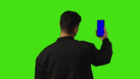 Rear-View-Of-Young-Man-Holding-Blue-Screen-Mobile-Phone-To-Camera-Standing-Against-Green-Screen