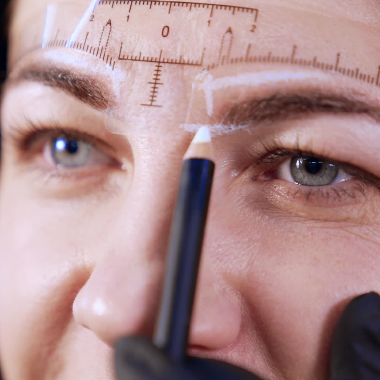 Upper part of the face of mid-aged woman with ruler attached to her forehead. Cosmetologist applies lines for permanent eyebrow make-up with white pencil. Close up