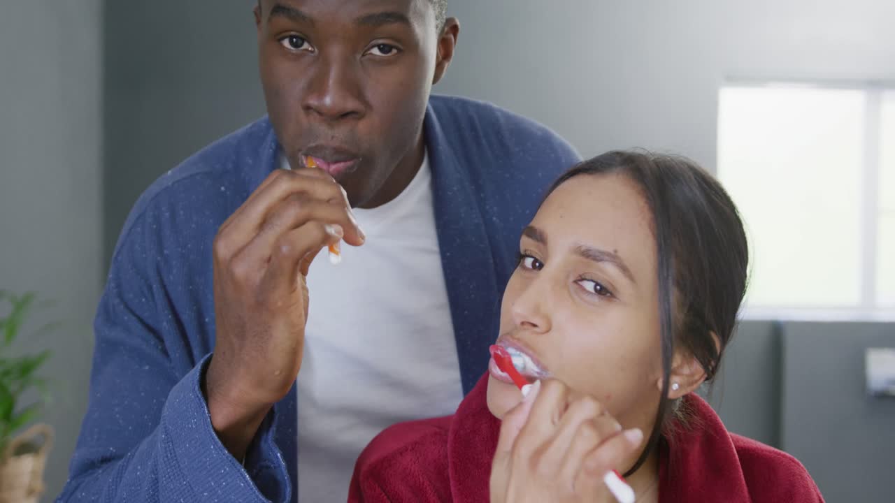 Video of happy diverse couple brushing teeth and smiling in bathroom at home