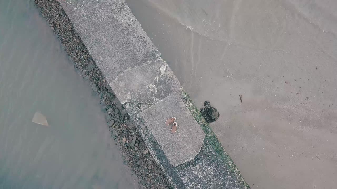 Shoes on seascape while rising tide gradually engulfing sandy shoreline near wooden pier during bright summer day