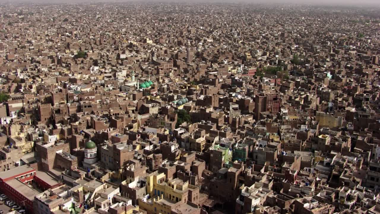 Drone footage captures the sprawling cityscape of Multan, Pakistan, showcasing a densely packed urban environment with low-rise buildings, narrow alleys, and scattered green domes