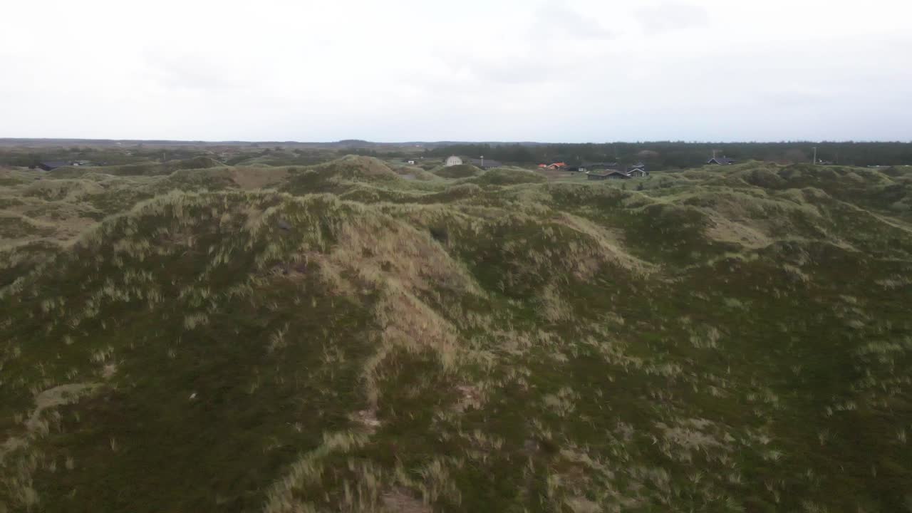 un dron orbita a la izquierda sobre las dunas en la playa de hvide sande, revelando casas anidadas en las dunas mientras el cielo gris y los vientos tormentosos barren el paisaje en una noche de mal humor.
