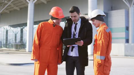 Investor-Des-Projekts-In-Einem-Schwarzen-Anzug,-Der-Das-Bauobjekt-Mit-Bauarbeitern-In-Orangefarbener-Uniform-Und-Helmen-Untersucht