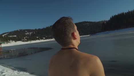 hombre sin camisa sentado en la nieve al lado de un lago congelado respirando pesadamente mientras prepara la mente y el cuerpo para sumergirse en agua helada