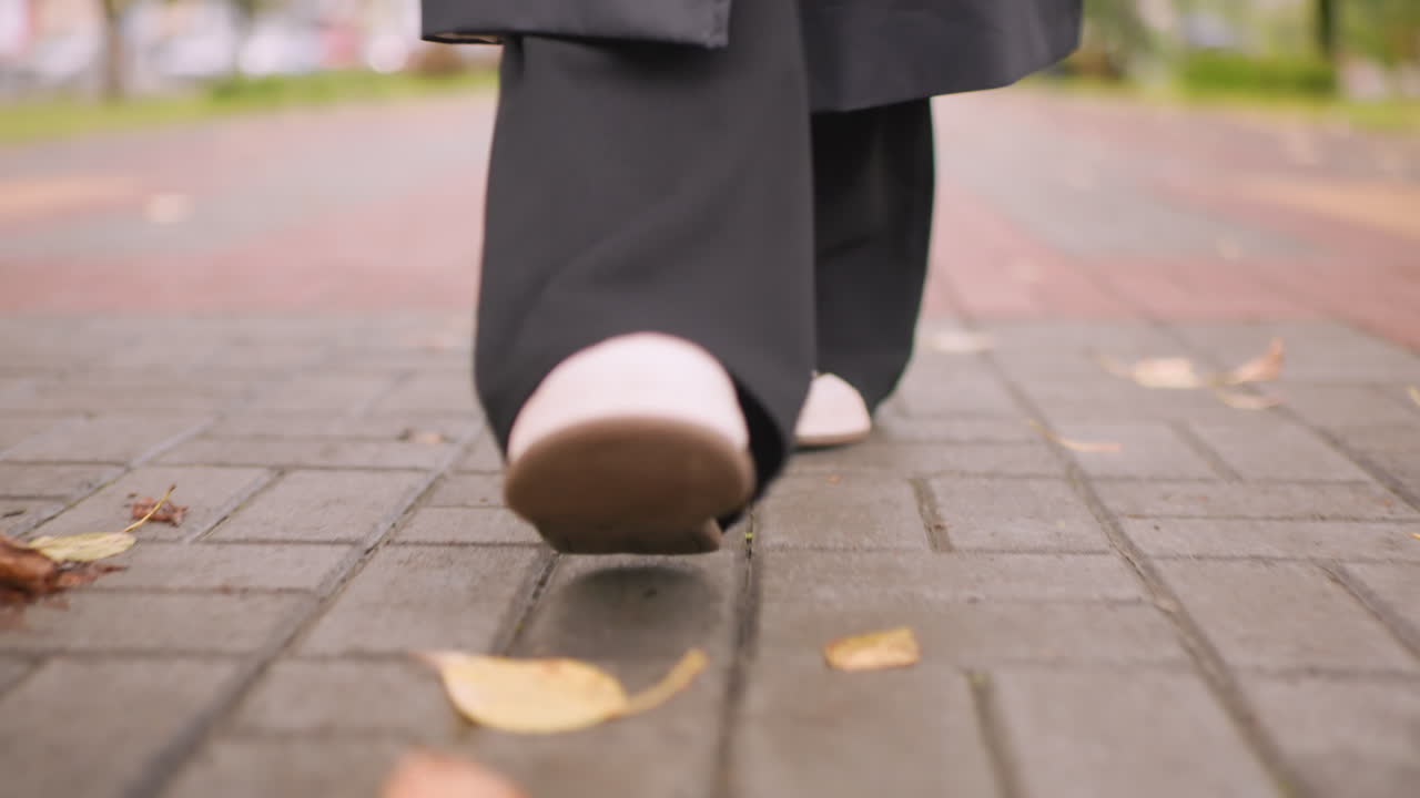 Close-up of woman walking on brick path wearing long black coat and white sneakers, autumn leaves scattered on ground, low angle view capturing detail of steps, urban outdoor atmosphere moment