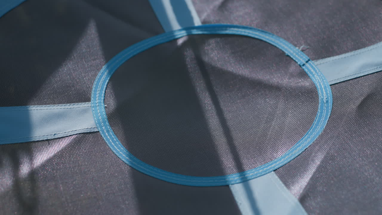 Close up of round blue swing seat featuring reinforced stitching around center mesh ring, illuminated by soft daylight, intersecting bands, and gentle shadow play from nearby frame