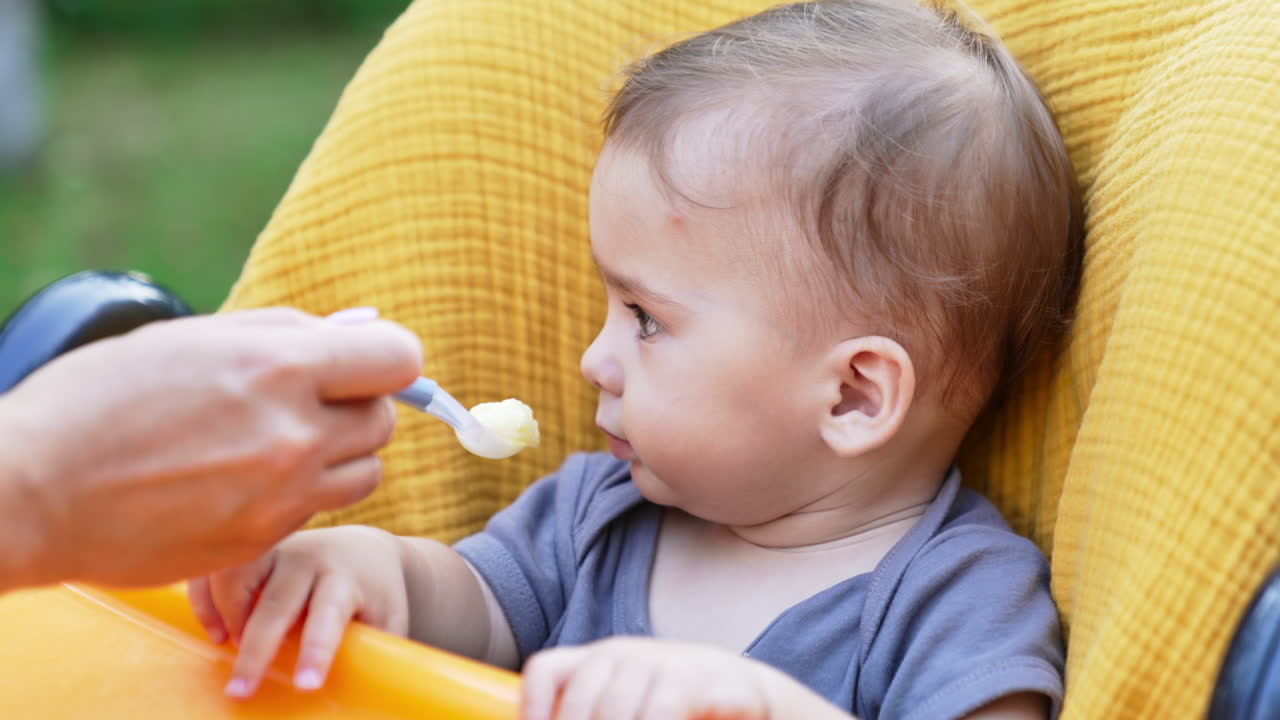 Beautiful Caucasian child refusing to eat. Mom's hand giving a spoon to her son but he turns his head away. Close up.