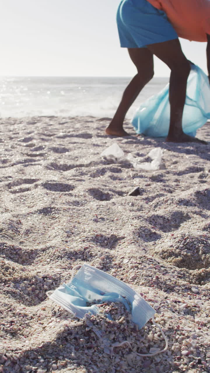 Video vertical de hombre afroamericano separando residuos en la playa.