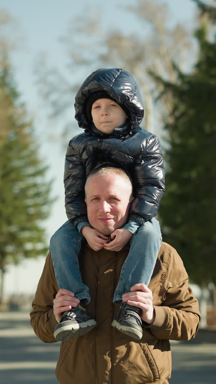 padre llevando a su hijo en sus hombros ambos sonriendo, el padre lleva una chaqueta marrón y gorra, y el niño está en una chaqueta negra con árboles y postes de luz detrás