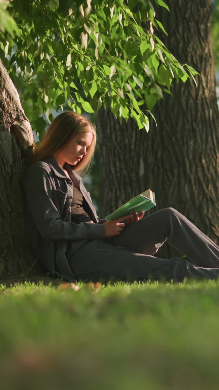 mujer sentada al aire libre con las piernas estiradas, apoyada en un árbol en un campo de hierba, leyendo un libro bajo la cálida luz del sol, las hojas de los árboles balanceándose suavemente en la brisa, con el fondo con vegetación