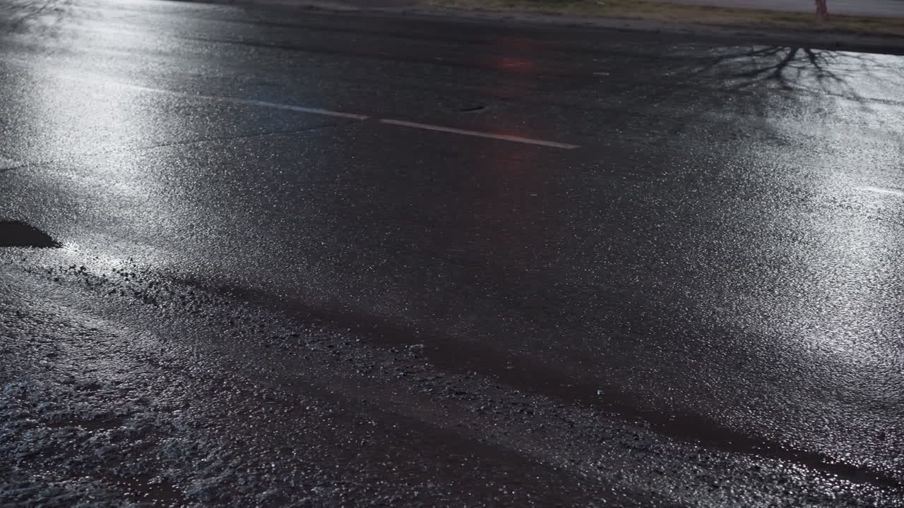 밤에 은 도로의 클로즈업 뷰, 표면에 빛의 반사와 도로에 나무 반사와 함께 지나가는 자동차를 보여줍니다.