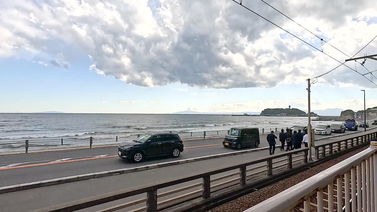 Vehicles and pedestrians move along a scenic coastal road in Kamakura, Japan under a partly cloudy sky