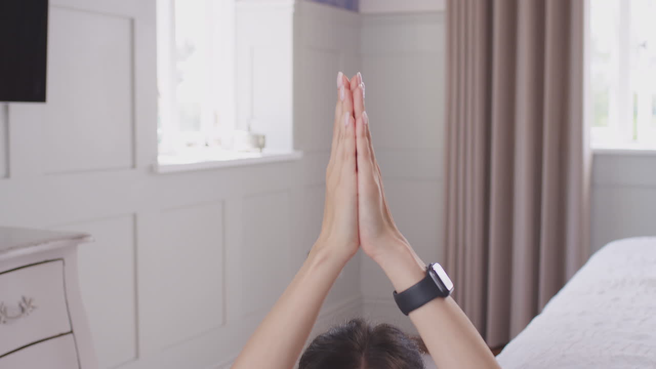Close up of woman's hands coming together in yoga pose before camera tilts down to show her sitting on exercise mat in bedroom - shot in slow motion