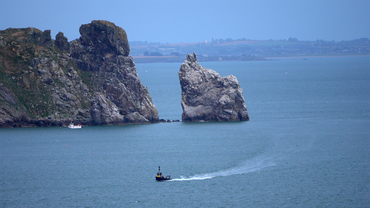 A motorboat speeds across the waters near Howth Peninsula, Dublin, leaving a white trail behind. Rugged cliffs rise in the background, contrasting with the deep blue sea.