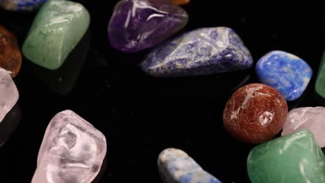 Vibrant chakra stones tumble onto a black surface, showcasing their colors and textures in slow motion under soft lighting
