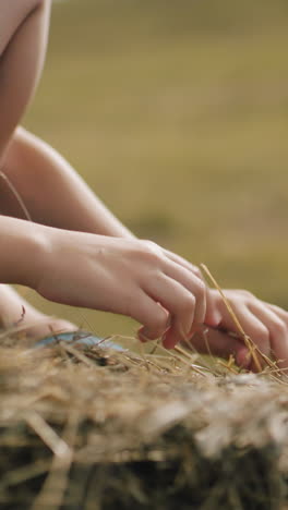 primer plano de las manos de un niño jugando con heno mientras está sentado en una bala de heno en un campo abierto, la luz del sol destaca las texturas suaves de la hierba seca, creando una atmósfera rural cálida y pacífica