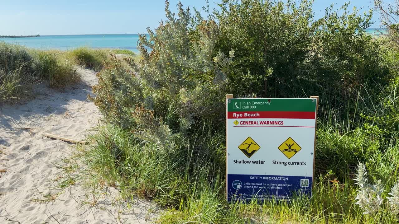 Static shot of beach warning sign, coastal vegetation, sand path, and ocean under daylight