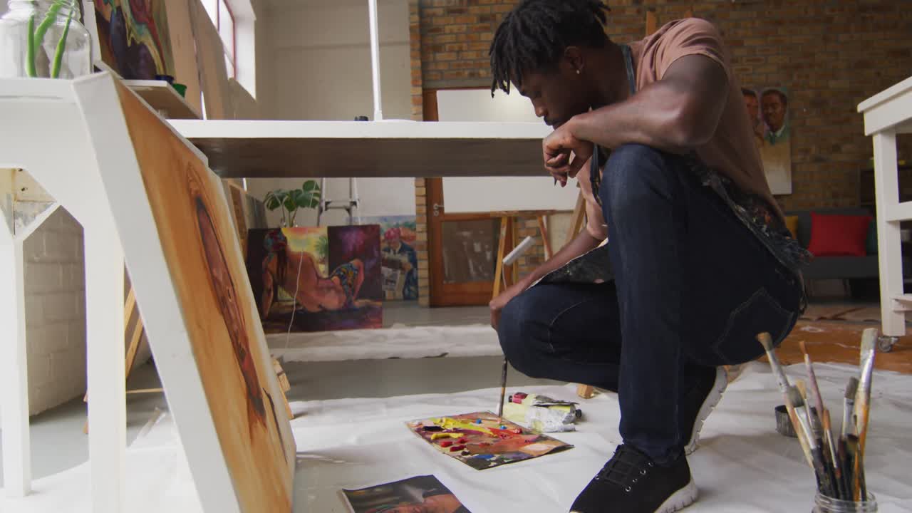 vista lateral de un artista masculino afroamericano con un delantal pintando en lienzo en un estudio de arte