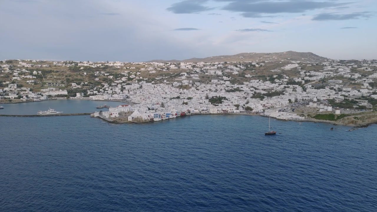 Derin mavi su üzerinde Mikonos'un uzak drone panoraması, açık denizde demirlemiş yalnız bir yelkenli tekne ve kıyı şeridi boyunca uzanan beyaz kasaba ile.