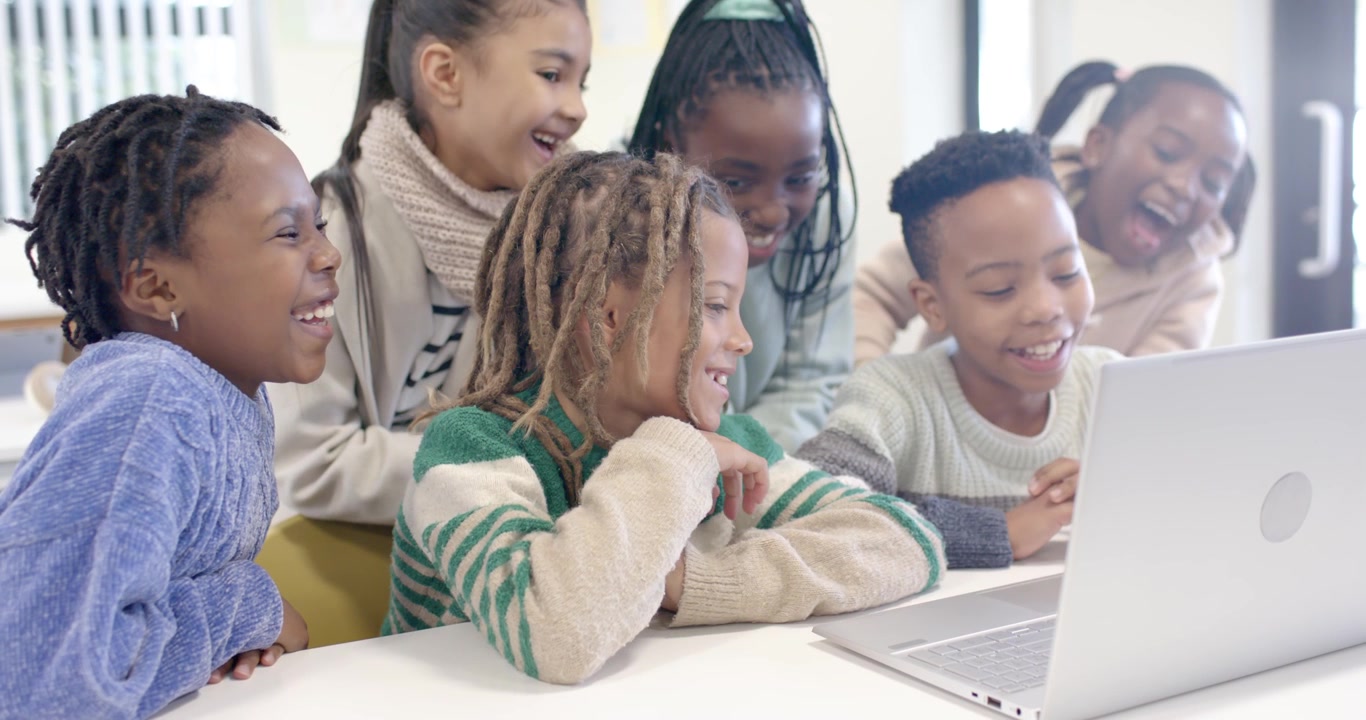 Preadolescenti afroamericani riuniti attorno a un laptop argentato su un tavolo, con un operatore che digita e reagisce a un video
