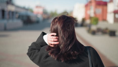 close-up back view of lady with brown hair in a black coat adjusting her head, showing off three rings on her left hand, on a sunny urban street