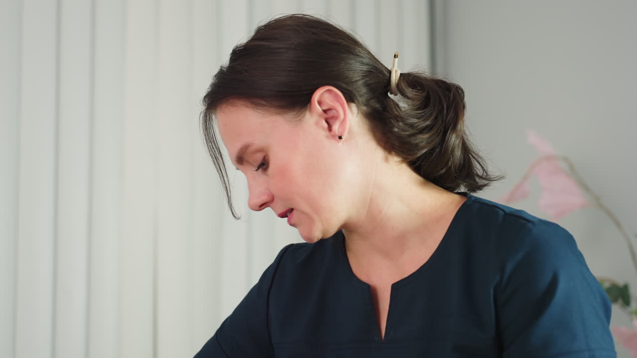 Portrait of fair skinned aesthetician in navy blue work dress focused while working showing professionalism and concentration in spa clinic environment with blinds background and pink plant decor