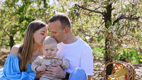 loving caucasian parents kiss their toddler baby on the blond head. man kisses his wife on the forehead. family together in the nature.