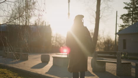 back shot of a woman in a brown coat, jeans, and white shoes, carrying a backpack as she walks through a park at sunset.the warm, golden light casts a soft glow, emphasizing the sadness in her posture