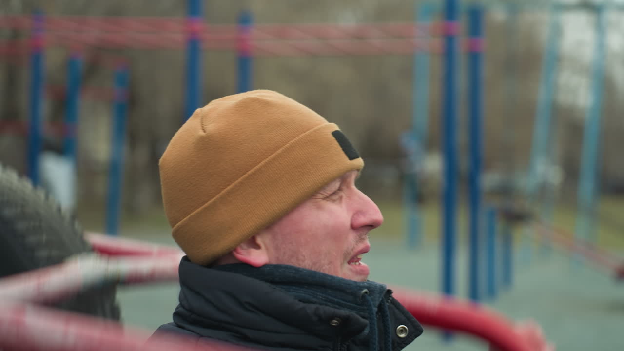 Close-up of a coach in a brown beanie doing pull-ups on workout equipment, pause to rest and looked left and then forward, with a blurred view of people in the background