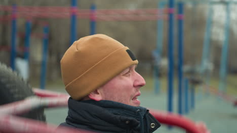 close-up of a coach in a brown beanie doing pull-ups on workout equipment, pause to rest and looked left and then forward, with a blurred view of people in the background