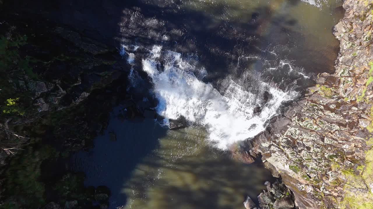 Drone footage captures cascading waterfall and faint rainbow over rocky gorge at Ebor Falls, New South Wales, under natural daylight with steady overhead perspective