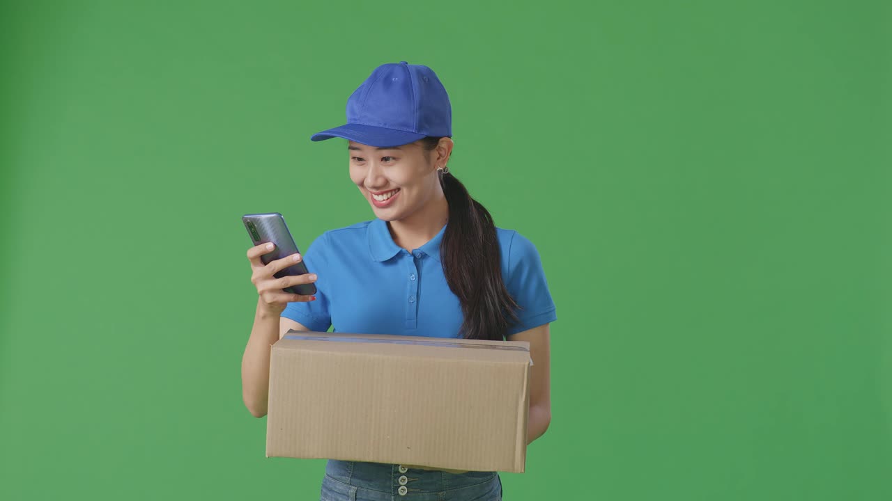 mensajera asiática en uniforme azul usando un teléfono inteligente mientras entrega una caja de cartón en el fondo de la pantalla verde en el estudio