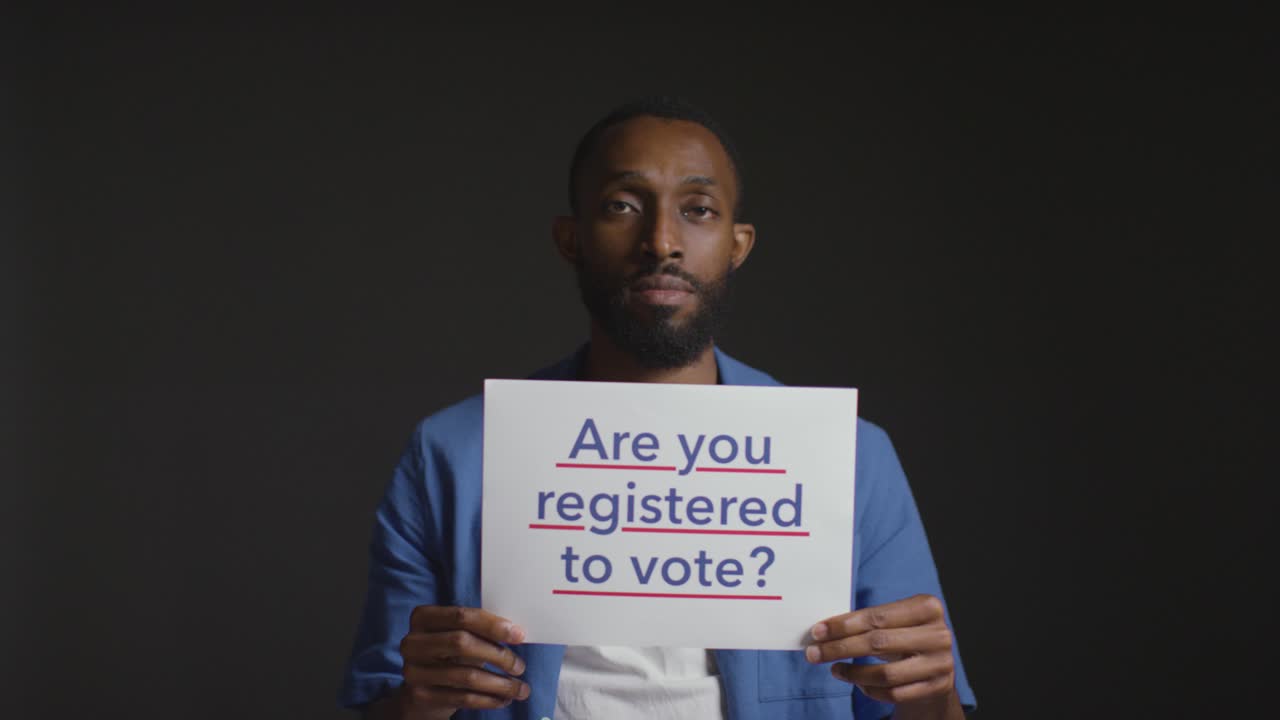Portrait Of Man Holding Are You Registered To Vote Sign In Election Against Black Background